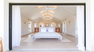 Vaulted primary bedroom with twin backlit mirrors on a grasscloth wall behind an upholstered bed at Cloudbreak, Barbados