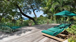 Wicker sun loungers with green cushions and a parasol on the timber deck, the sea beyond the trees, Fustic House in Barbados
