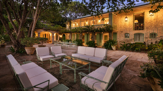Great House lit at dusk beyond a courtyard of white-cushioned sofas and a glass table at Fustic House, Barbados