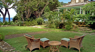 Wicker armchairs and footstools around a round table on the Fustic House lawn below a green-trimmed veranda, Barbados