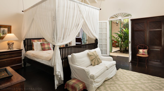 Dark wood bed under a sheer canopy, with a white loveseat and doors to the atrium, at Fustic House in Barbados