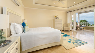 Cream tufted bed and blue floral rug in a bedroom at Monkey Hill, Barbados, with doors open to a sea-view balcony