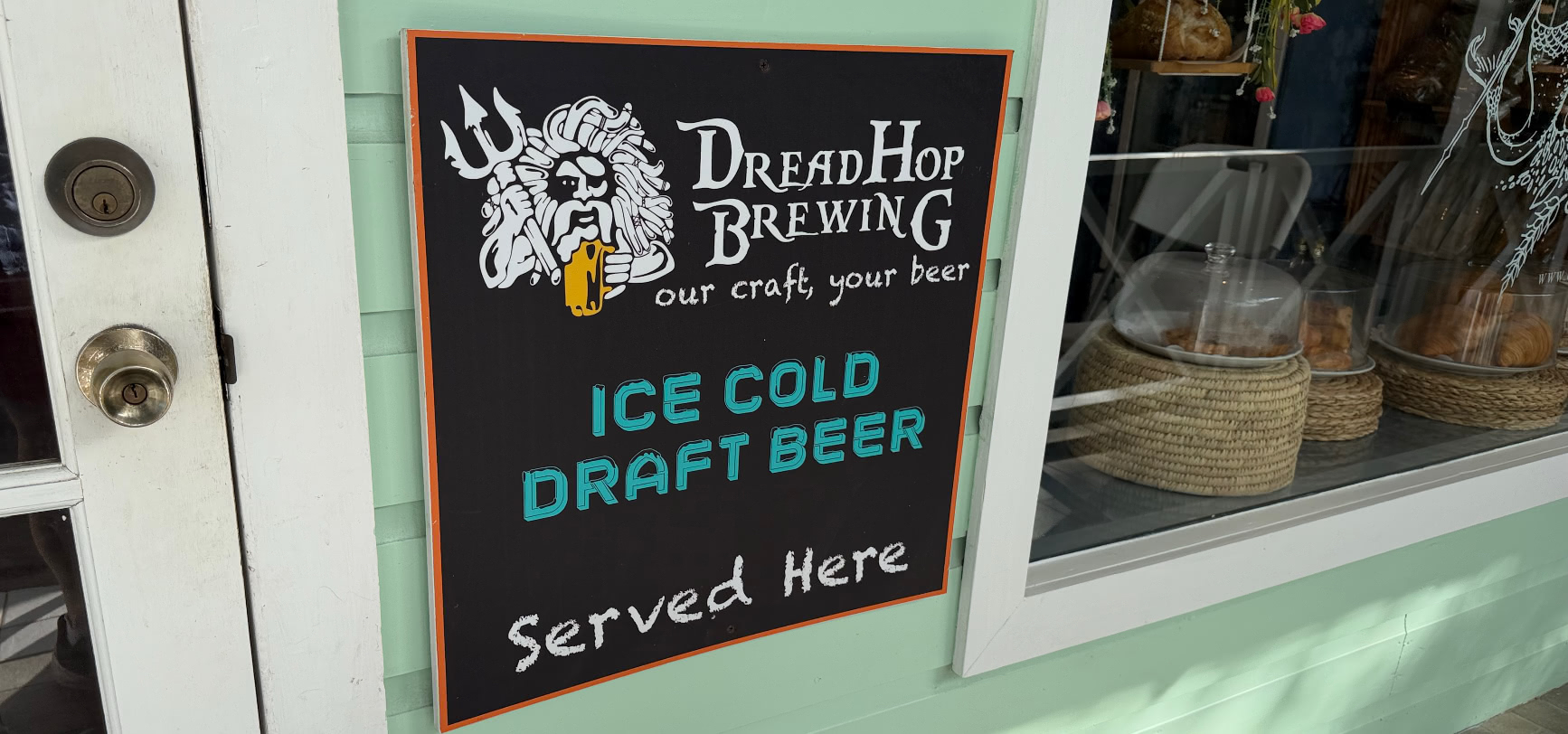A DreadHop Brewing 'ice cold draft beer served here' chalkboard by the door of The Cliff Bakery, added in 2025