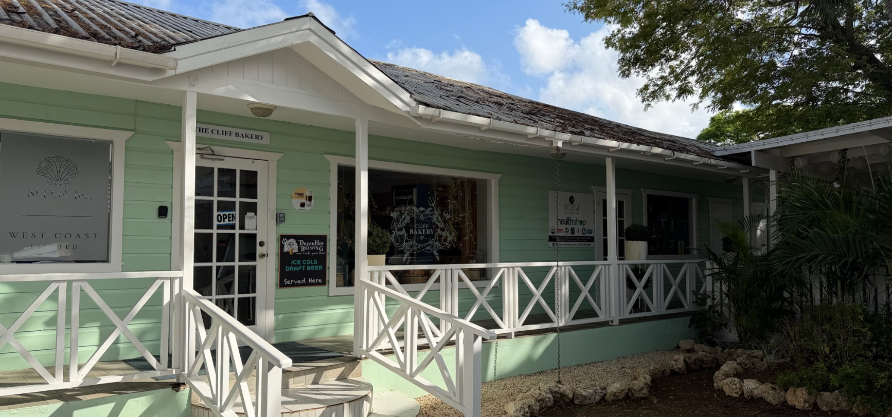 The sage-green clapboard shopfront of The Cliff Bakery in Holetown, Barbados, with white porch railings and an open sign