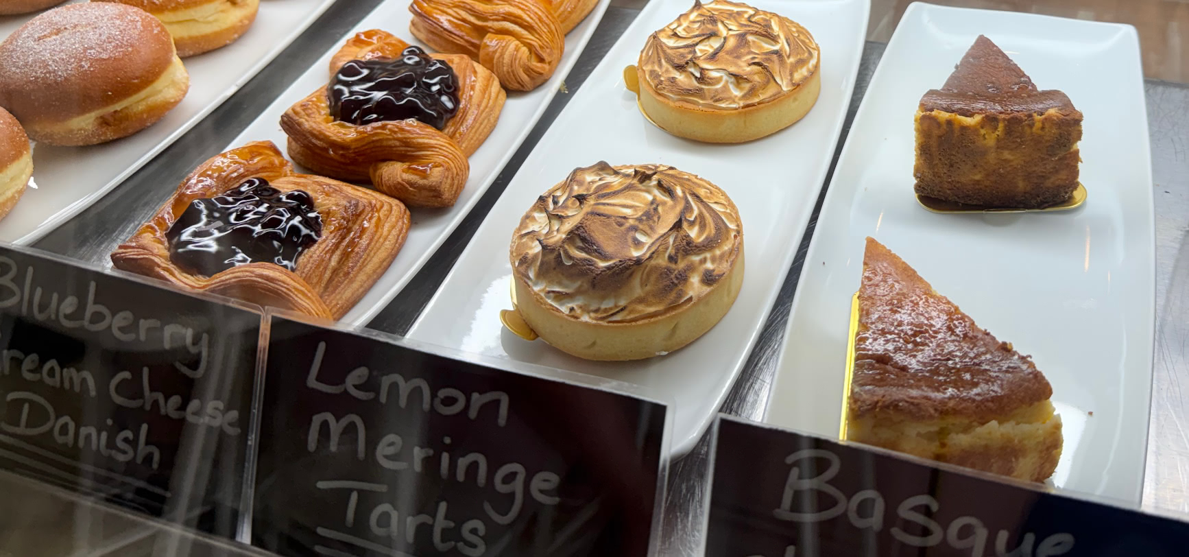 The chalk-labelled display case at The Cliff Bakery, with beignets, a blueberry cream cheese danish, lemon meringue tarts and Basque cheesecake