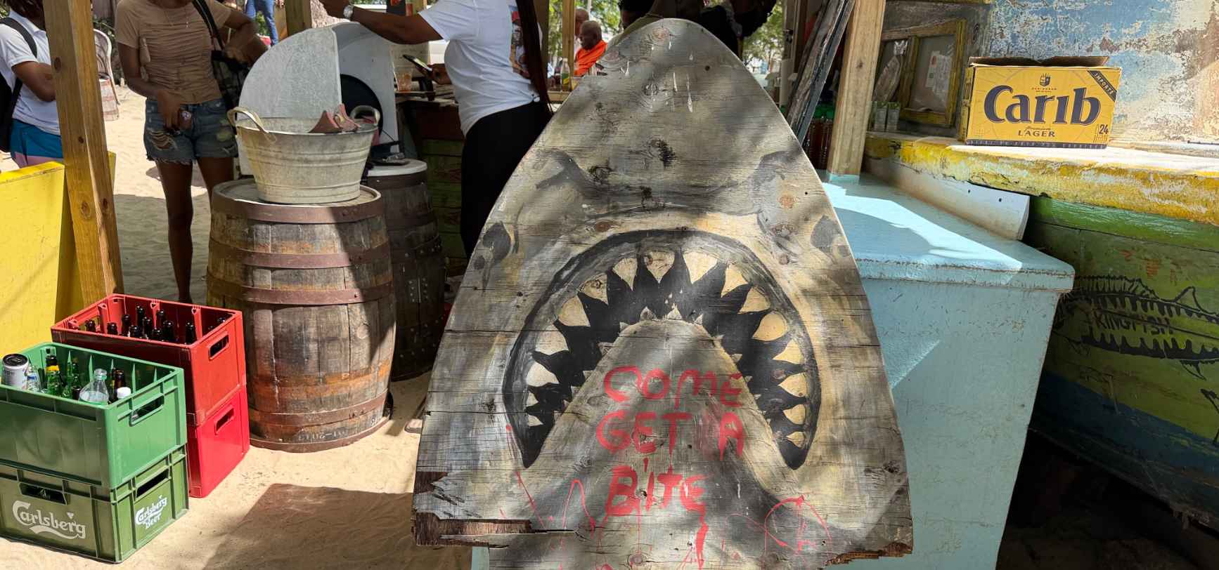 The hand-painted wooden shark-jaw sign at Caboose with teeth and red lettering reading COME GET A BITE, propped beside drinks crates