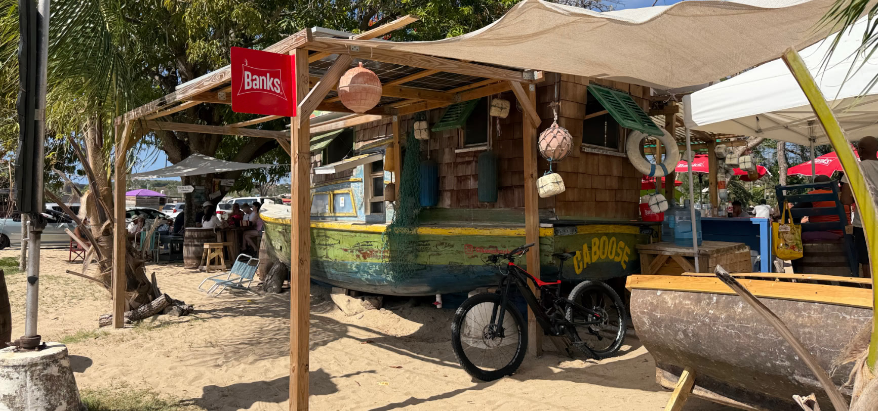 Side view of the beached wooden boat at Caboose with CABOOSE painted on the green hull, a red Banks sign and a timber awning