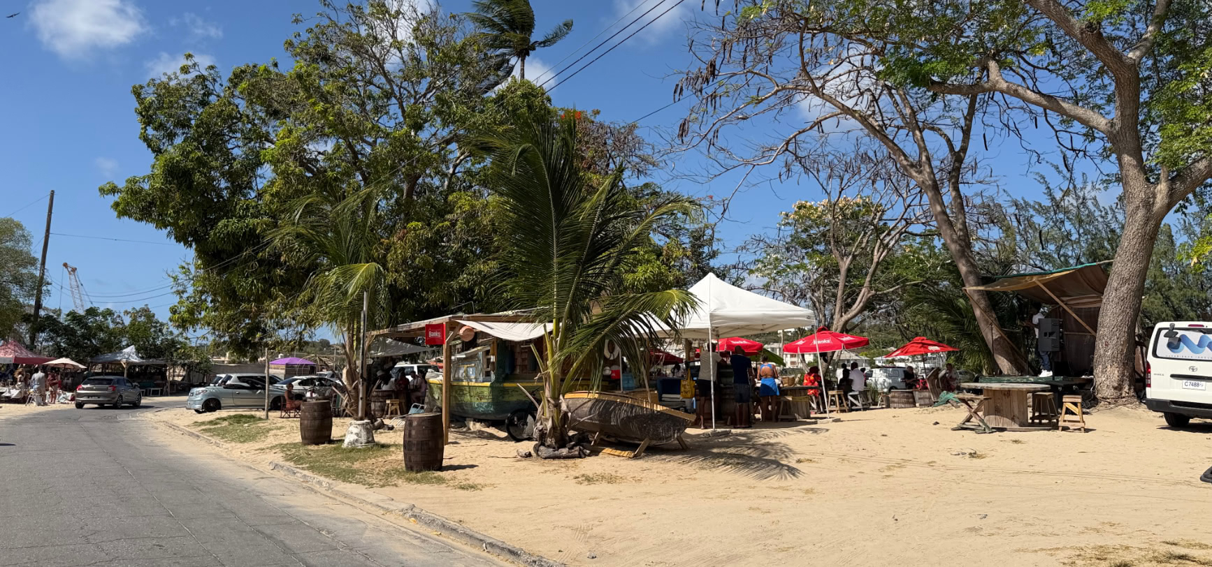 The sandy roadside lot at Caboose on the edge of Speightstown, with a white marquee, red umbrellas, big shade trees and a parked van