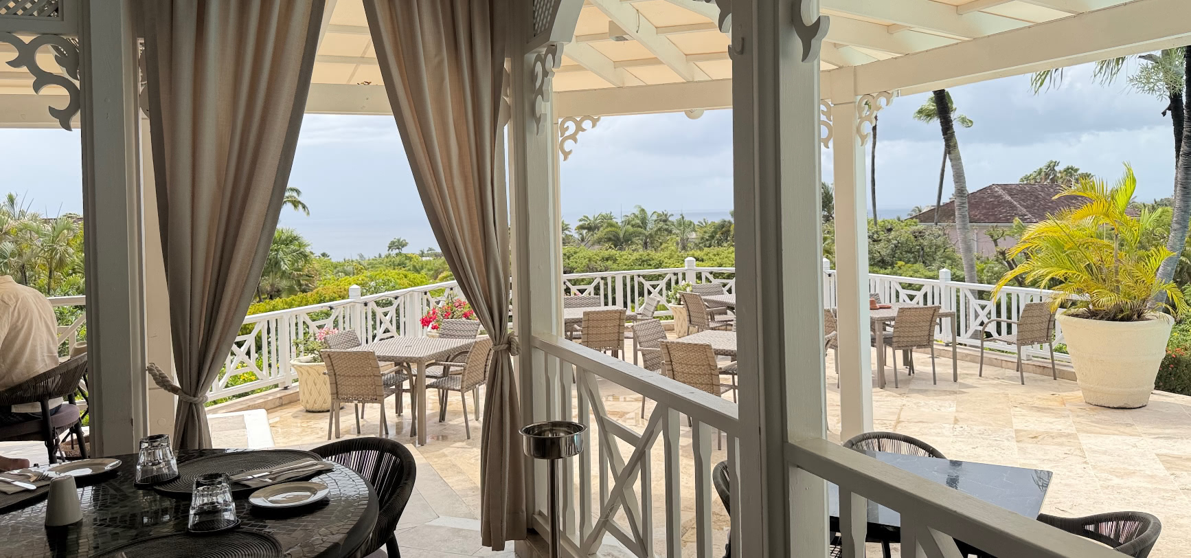 Covered terrace dining at The Deck, Sugar Hill, with white fretwork and drapes framing a view over the palms to the sea, Barbados