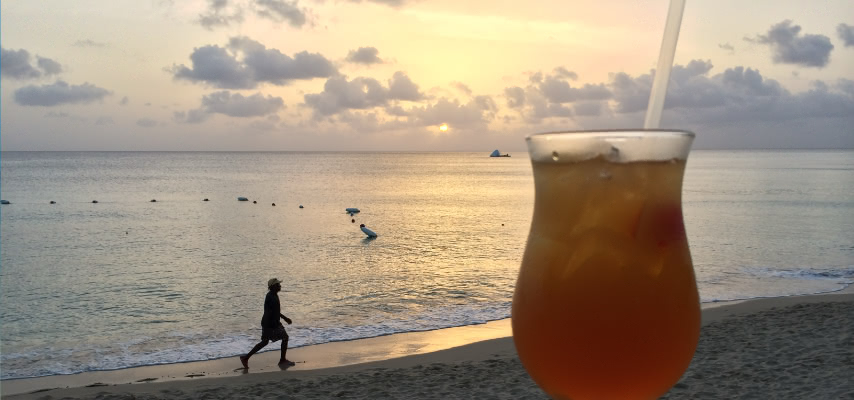 Sunset over a west coast beach in Barbados, with a sundowner on a beachfront table and a walker on the sand