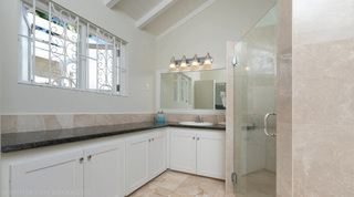 Bathroom with a long white vanity, scroll-grilled window and glass shower at Queens Fort Pavilion, Barbados