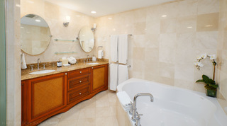 Twin-basin vanity, oval mirrors and corner bath in a cream-tiled bathroom, St. Peter's Bay 104 holiday apartment, Barbados