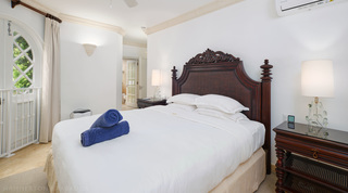 At St. Peter's Bay 308, Barbados, a carved bed with rolled blue towels, a woven-shelf bedside table and the bathroom beyond
