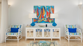 At St. Peter's Bay 401 in Barbados, white armchairs with striped cushions flank a mirrored sideboard and boat painting